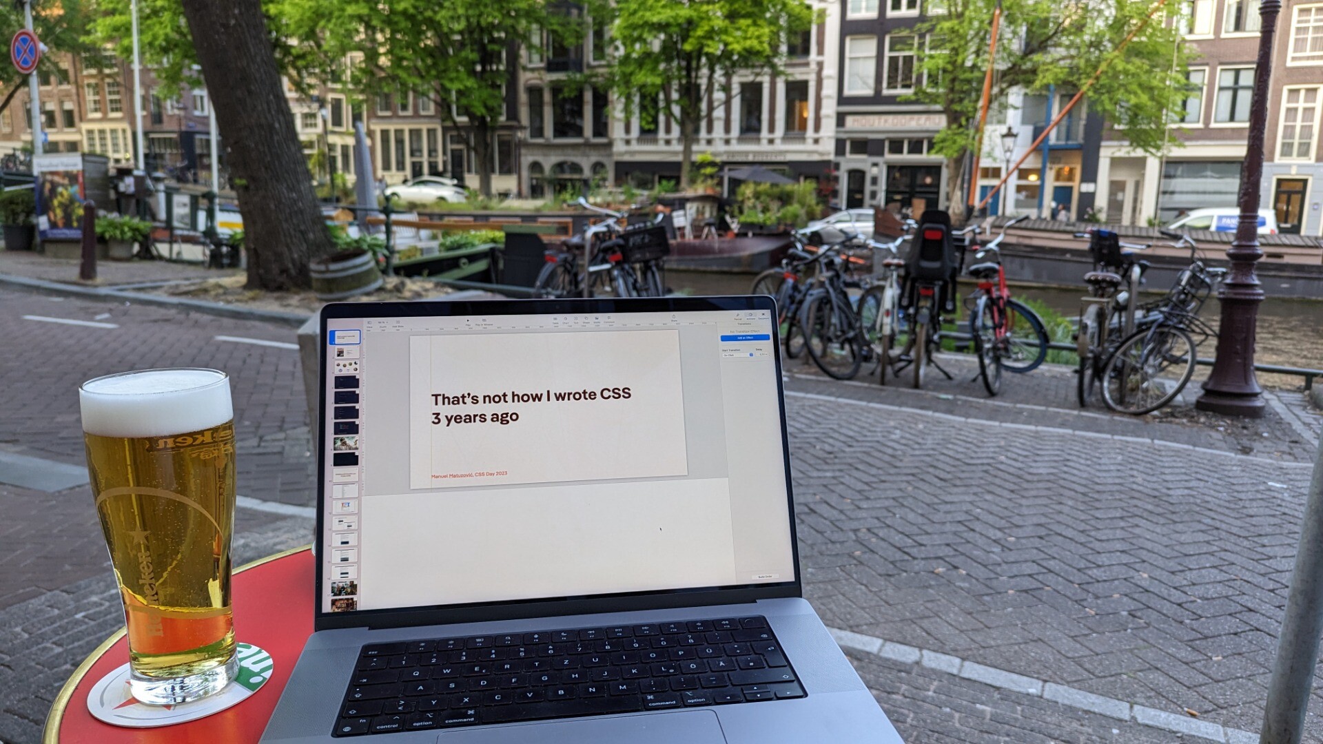 A beer and laptop on a small table in front of some bikes with a canal in the back, slide visible on the computer says: That's not how I wrote CSS 3 years ago
